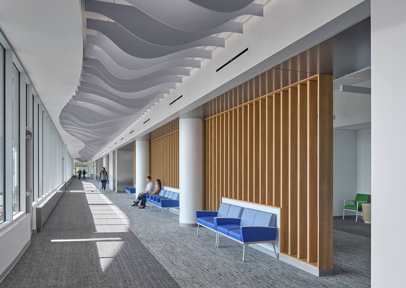 Modern building hallway with wavy ceiling, blue seating, and large windows.