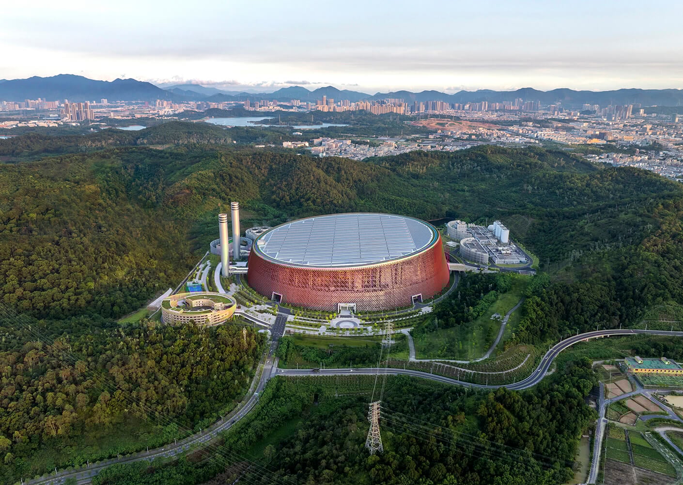 Aerial view of a modern circular building surrounded by lush greenery and distant cityscape.
