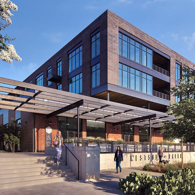 Modern office building with people outside, clear sky, and greenery around.