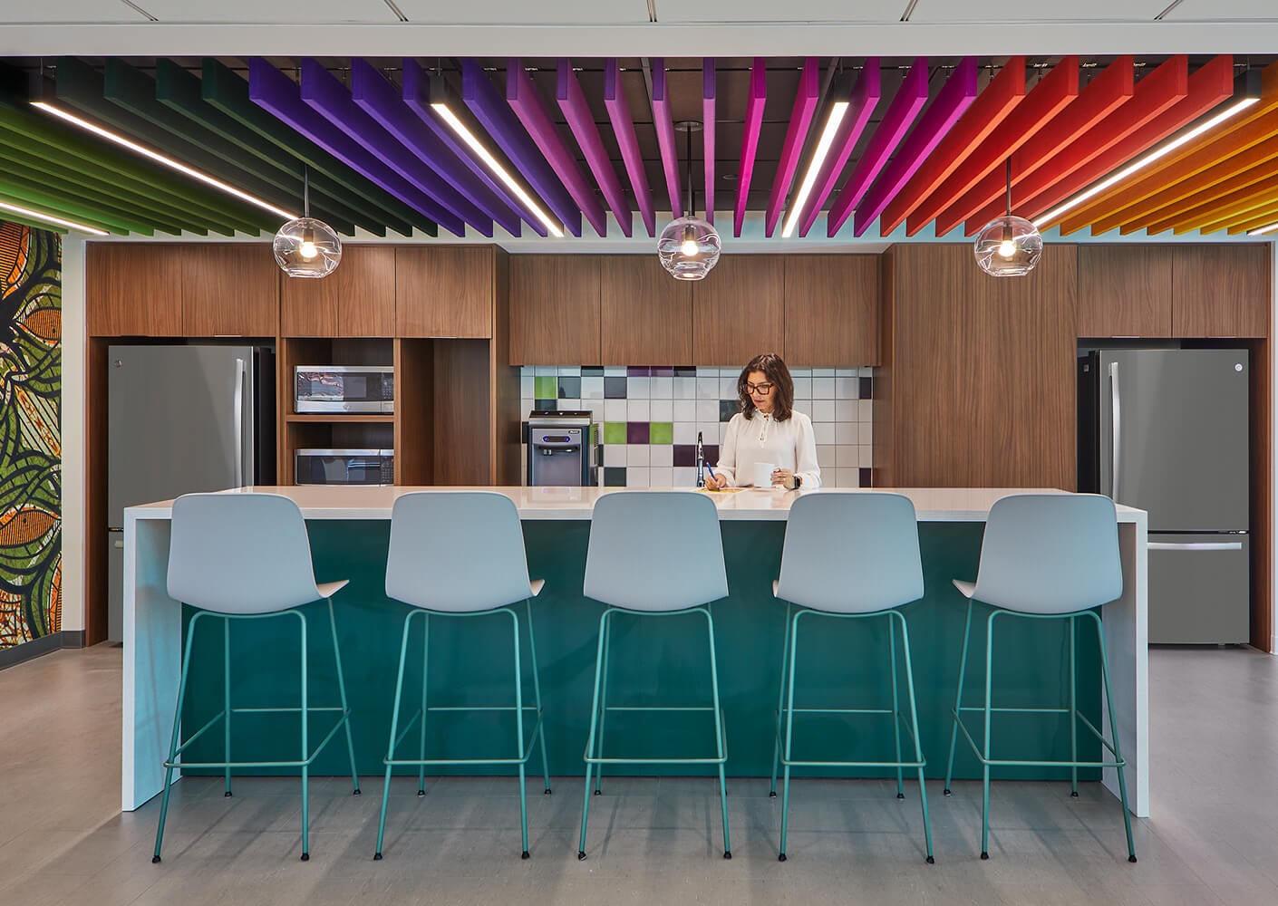 Modern office kitchen with colorful ceiling and woman standing at counter.