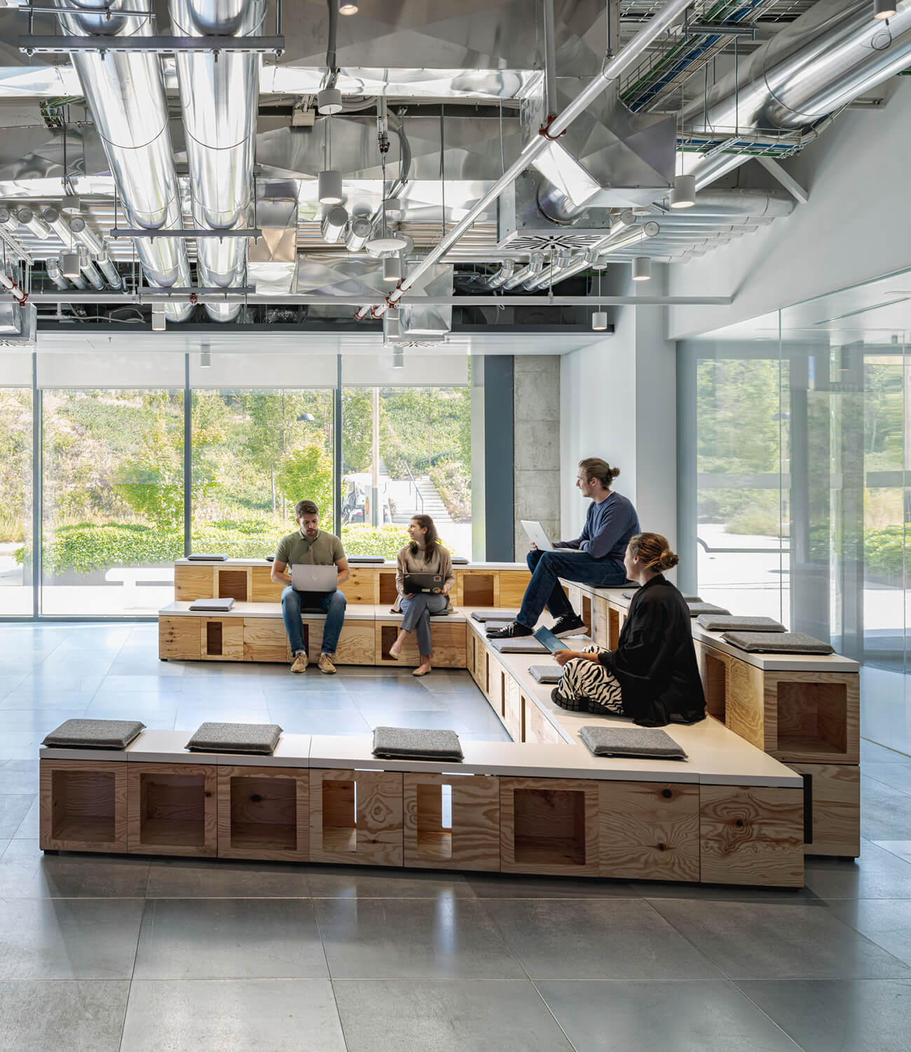 Modern office lounge with people sitting on wooden benches, working on laptops.