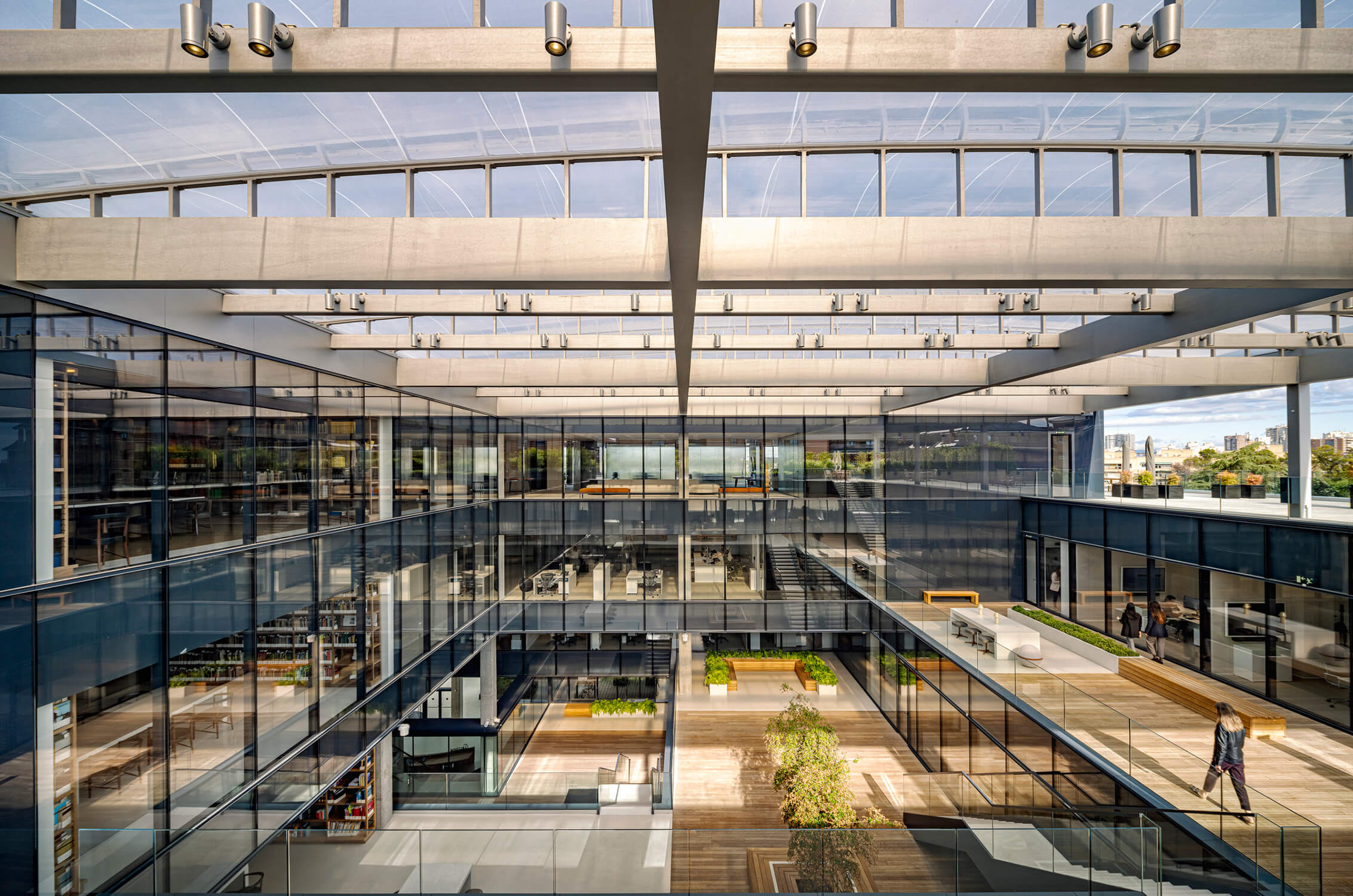 Modern office atrium with glass walls and open spaces, featuring greenery and seating areas.