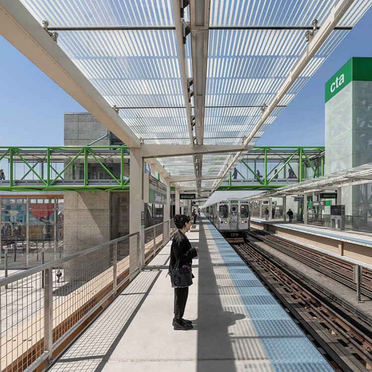 A person waits on the train platform as a train approaches with other commuters in the background