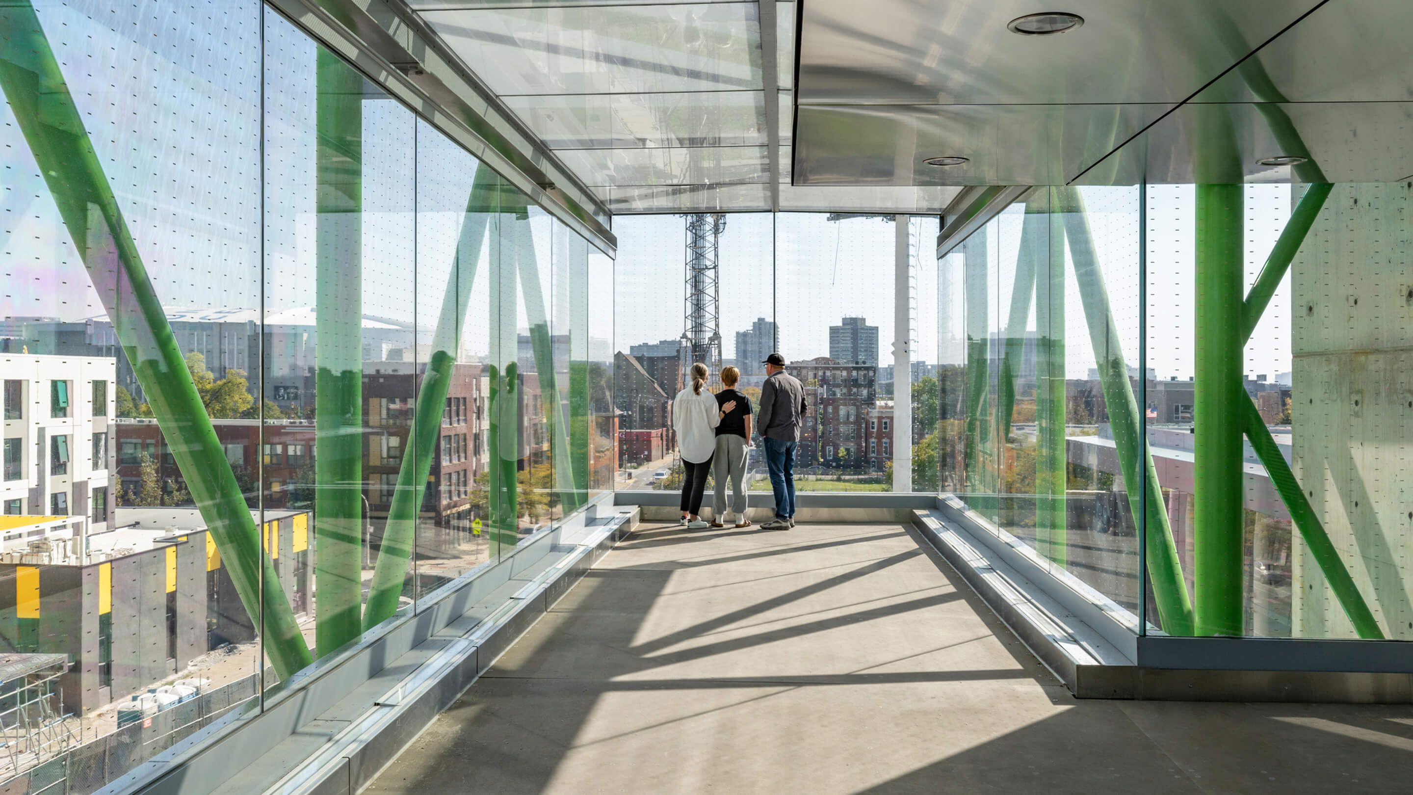 Modern walkway with glass walls, three people view cityscape from inside.