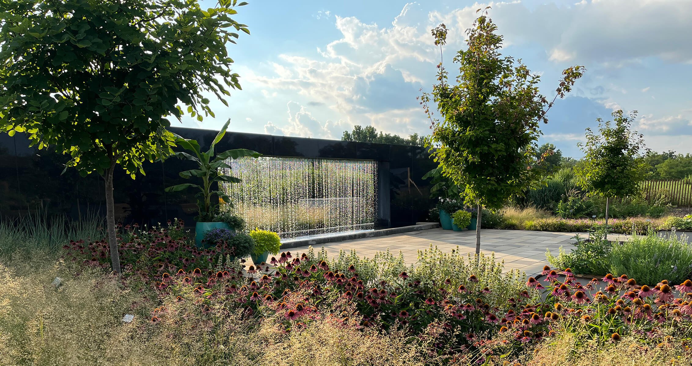 Garden with trees, vibrant flowers, and a cascading water feature under a blue sky.