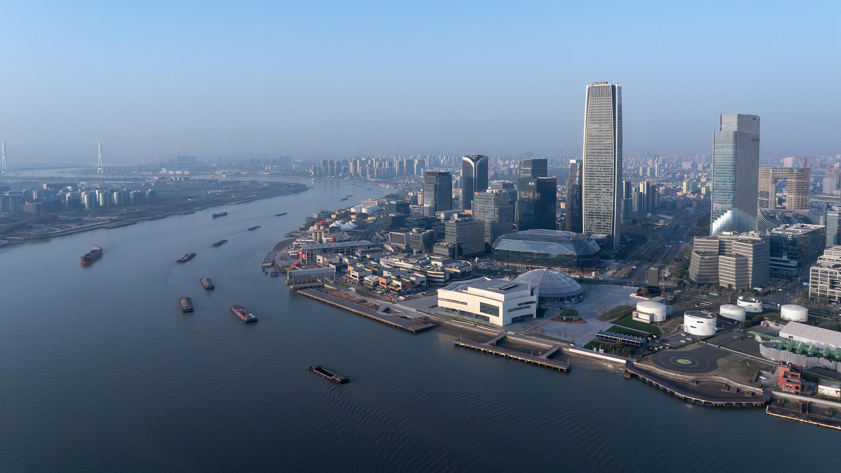 Aerial view of modern city skyline along a river at dusk with skyscrapers and cargo ships.