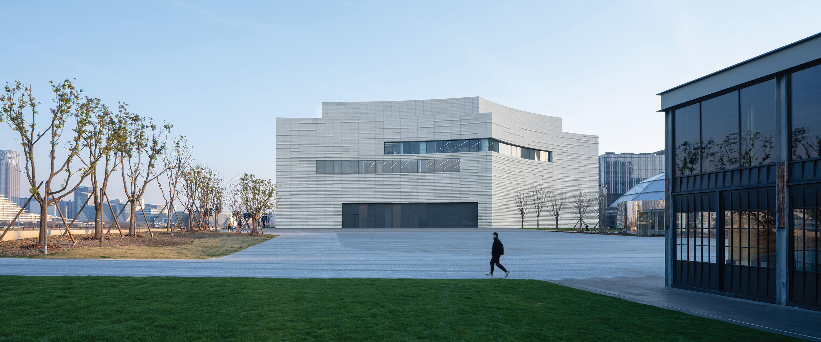 Modern white building with curves and angles, set behind a paved plaza and grassy area on a winter day.