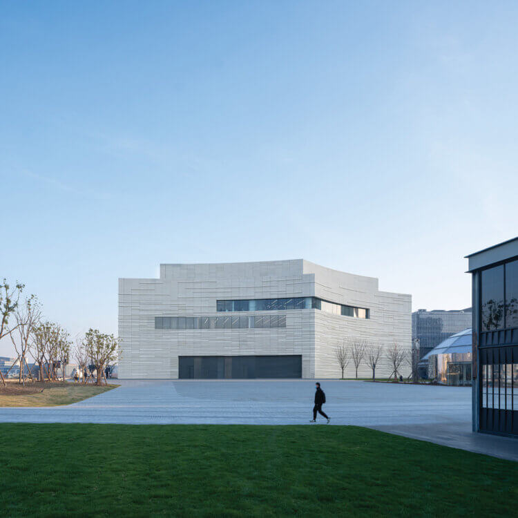 Modern white building with curves and angles, set behind a paved plaza and grassy area on a winter day.