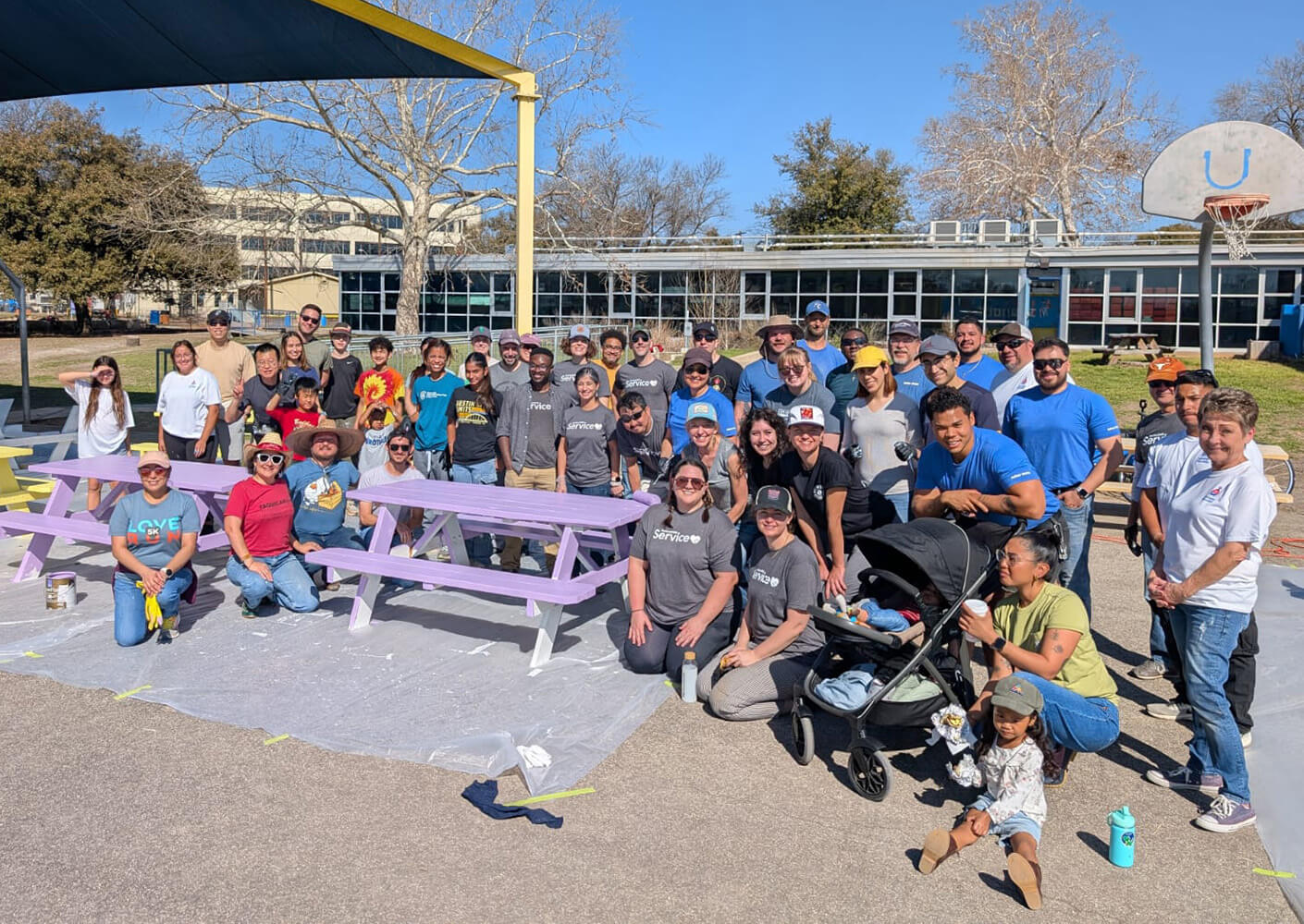 A group volunteering outdoors, painting purple picnic tables on a sunny day.