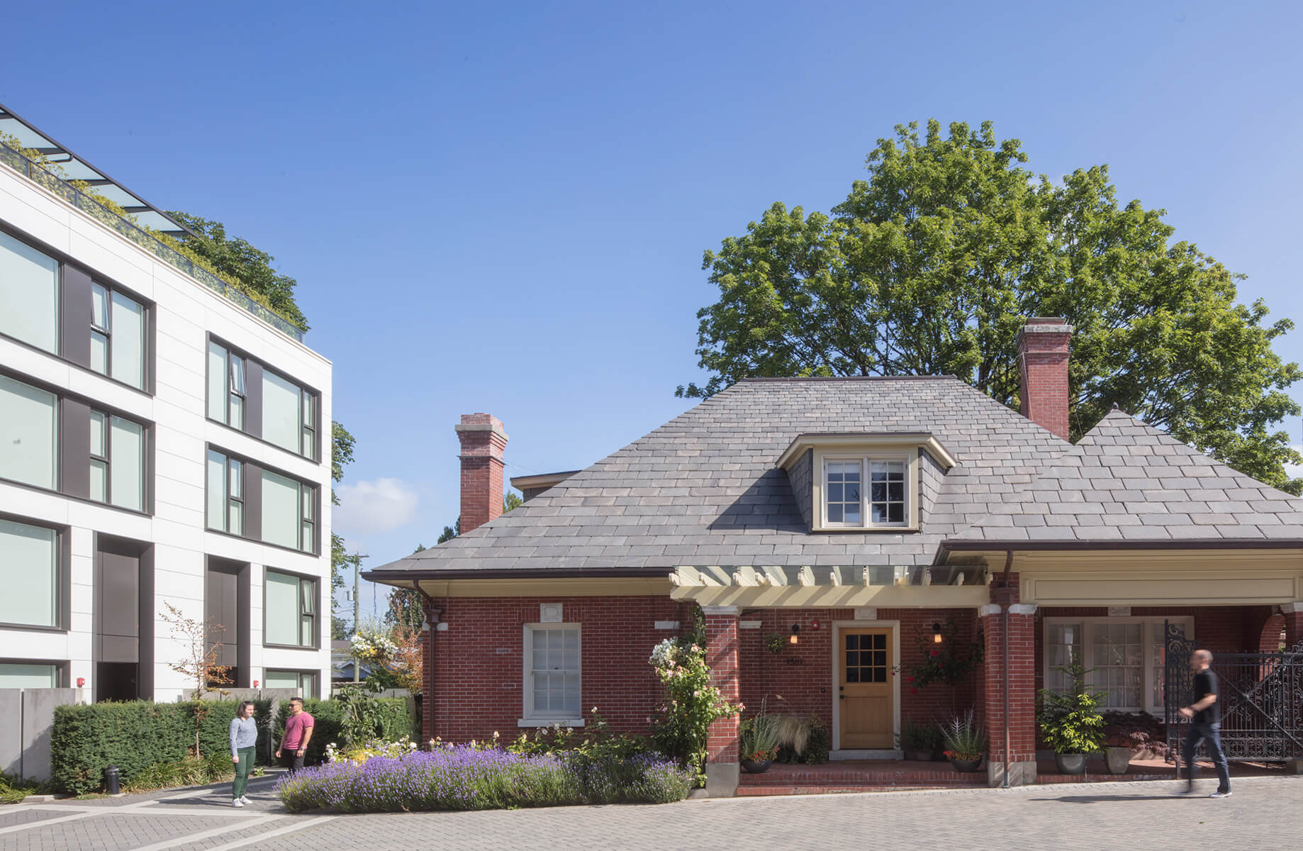Modern building next to traditional brick house with garden, under clear blue sky.