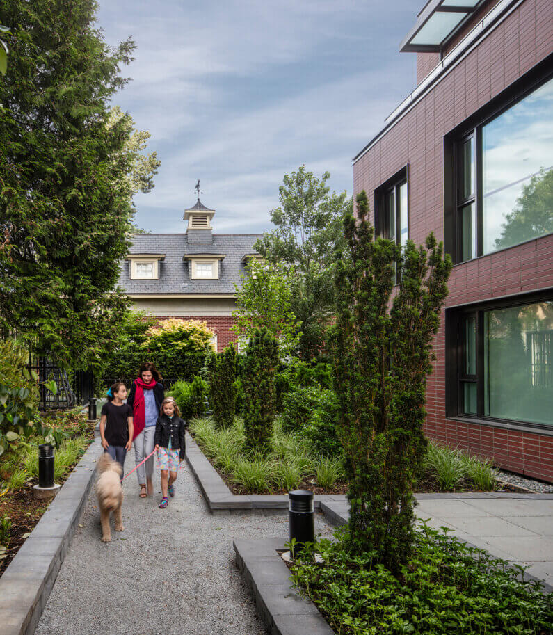 Family walking dog in a lush garden path beside modern building.