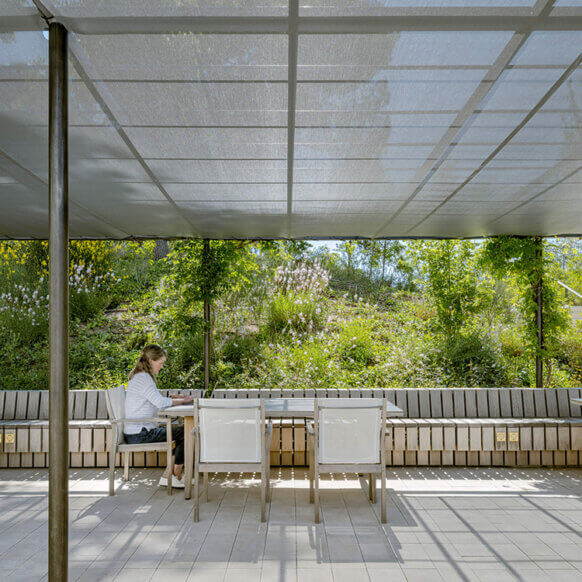 A person works at a table under a pergola in a lush garden setting.