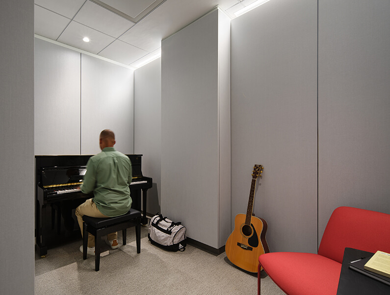 Man playing piano in a music practice room with guitar and chair nearby.
