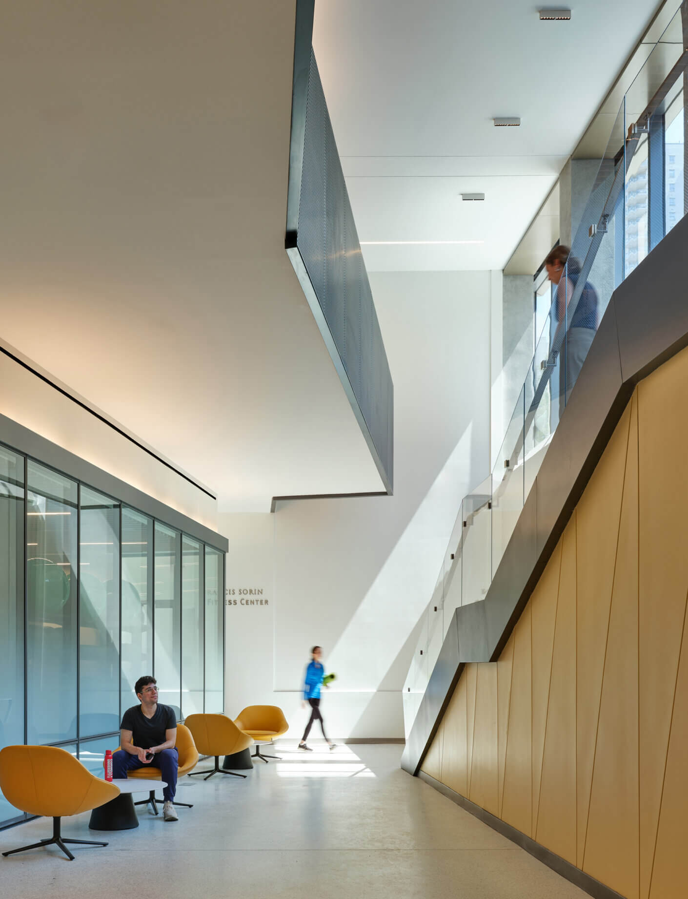Residential fitness lobby with glass, stairs, seating, and people walking in bright natural light.
