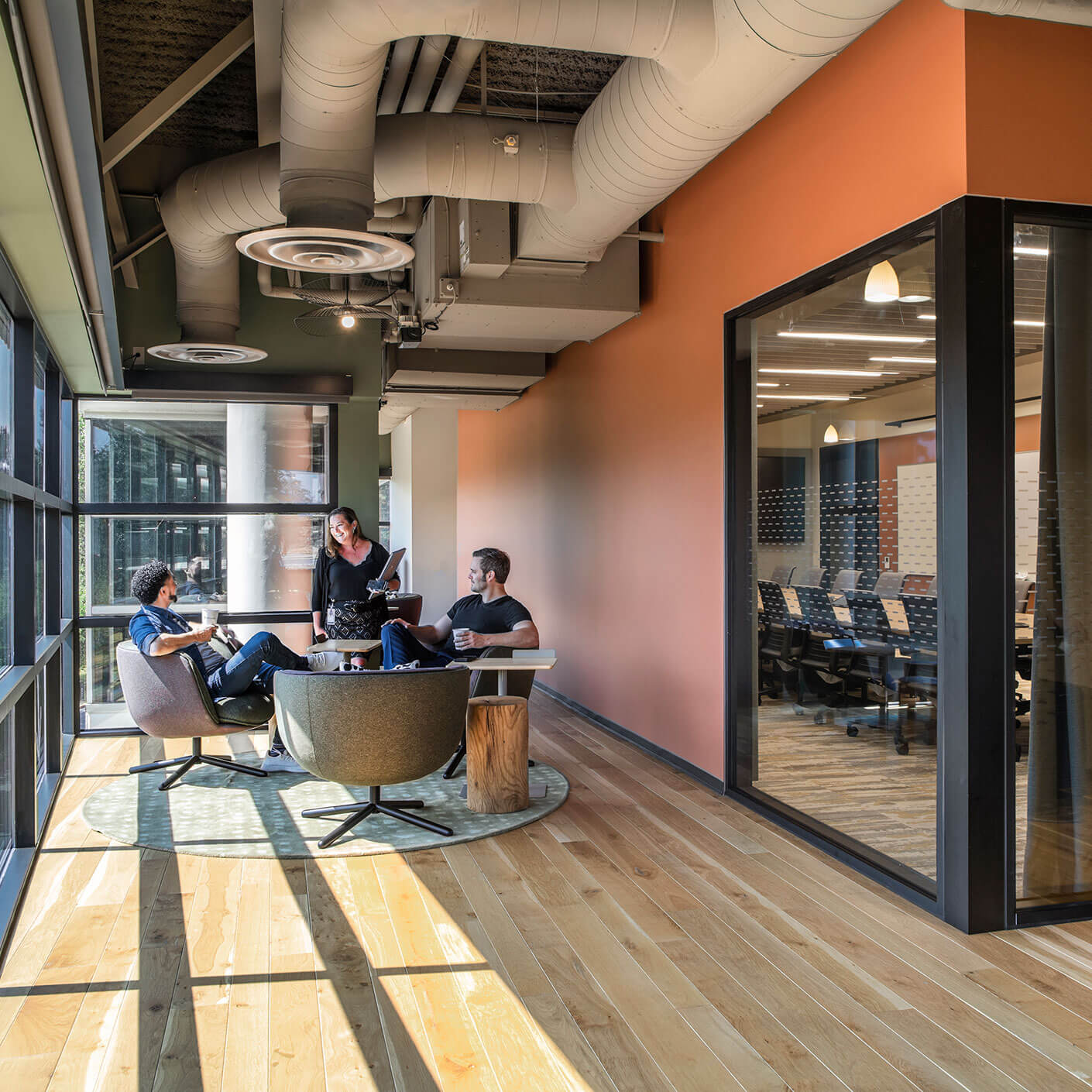 Modern office lounge with three colleagues conversing and a meeting room view.