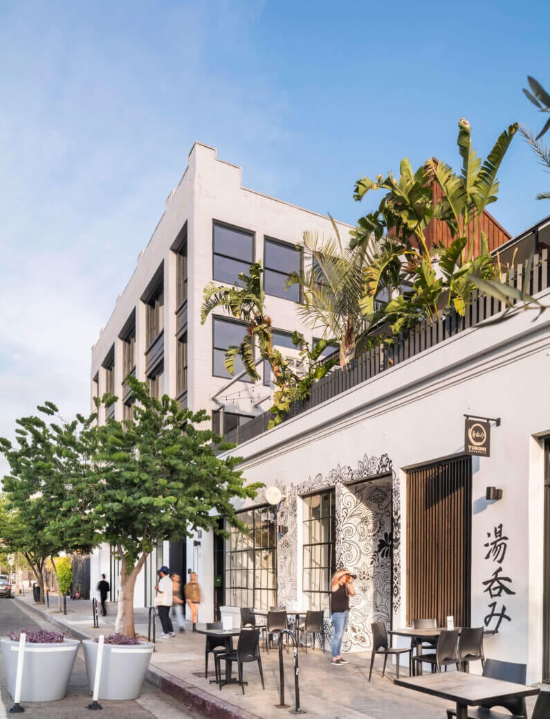 Street view of modern building with outdoor seating and tropical plants.