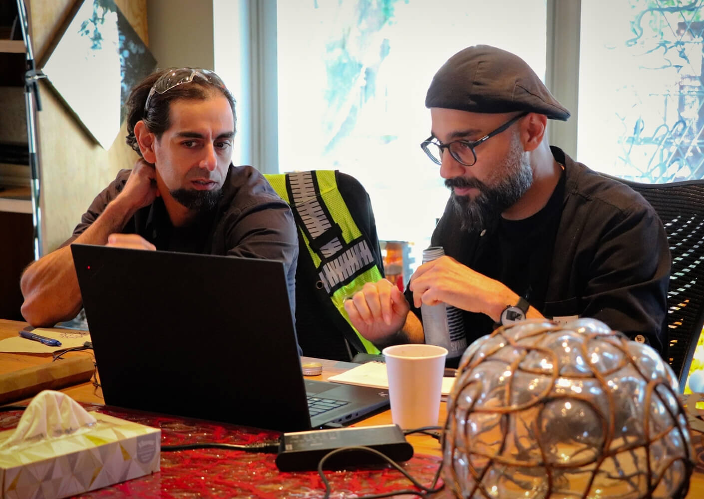 Two men discussing over a laptop at a table with various items, including a cup and tissues.