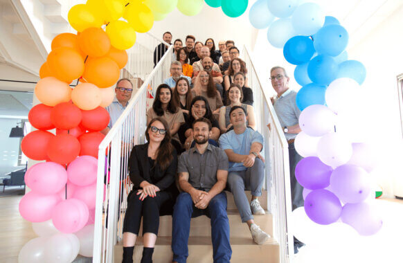 Team photo of diverse colleagues smiling on a staircase decorated with colorful balloons. 