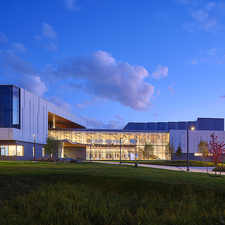 Modern architectural building with glass facade illuminated at dusk, surrounded by greenery.