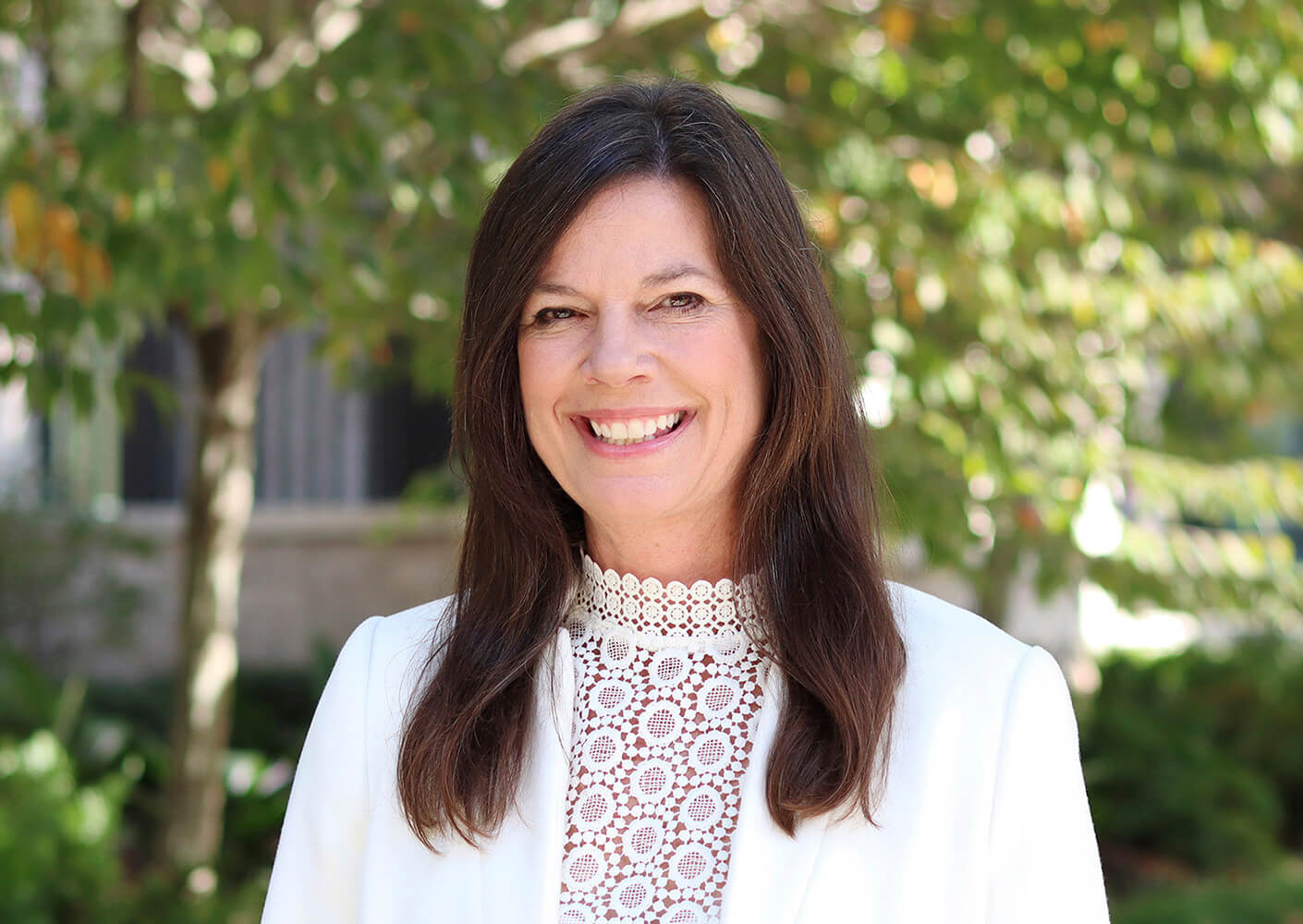 Smiling woman in a white blouse outdoors with greenery in the background.