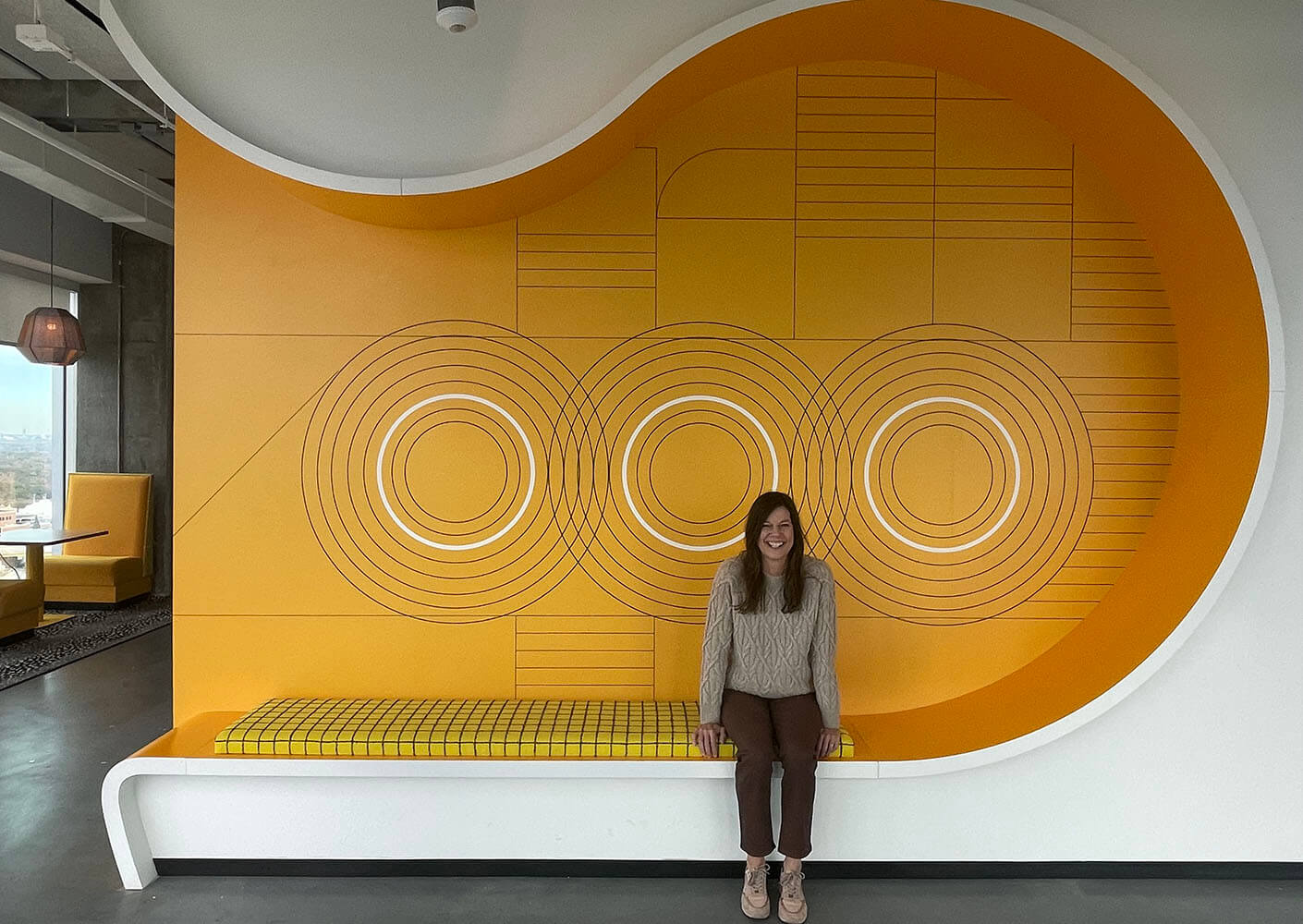 Woman smiling on a yellow geometric wall with circular designs in a modern interior.