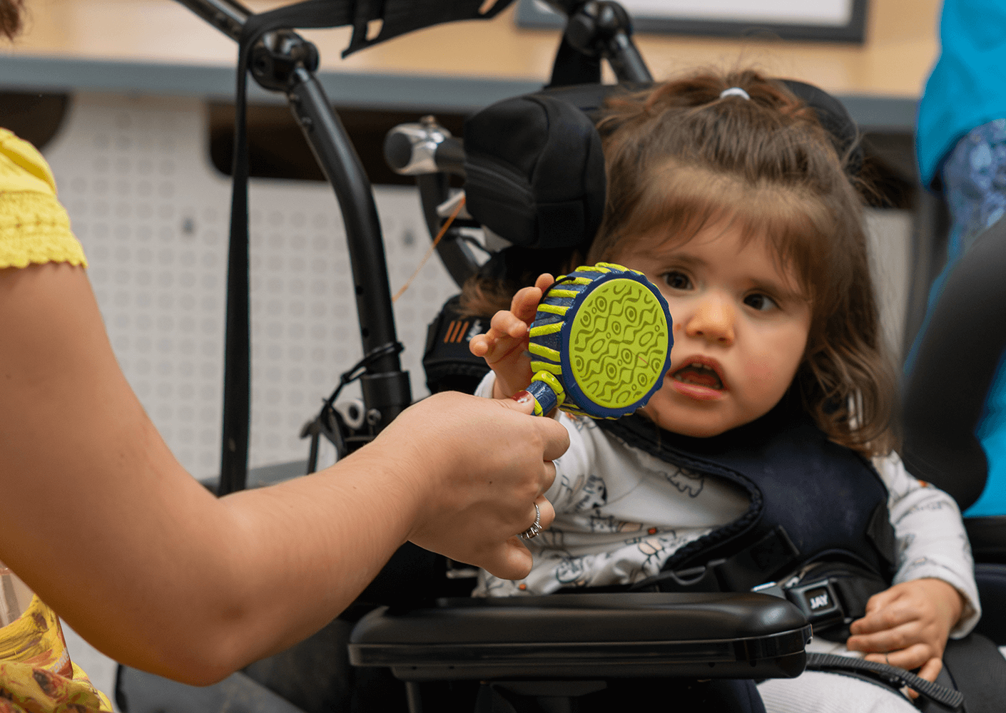 Child in a stroller reaching for a colorful toy with a caregiver nearby.