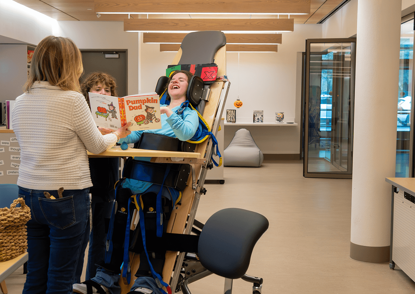 Child in supportive chair enjoying storytime with adults in a bright, accessible space.