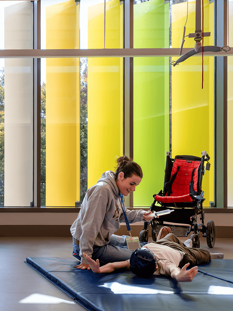 Therapist assisting a child in a therapy room with colorful windows in the background.