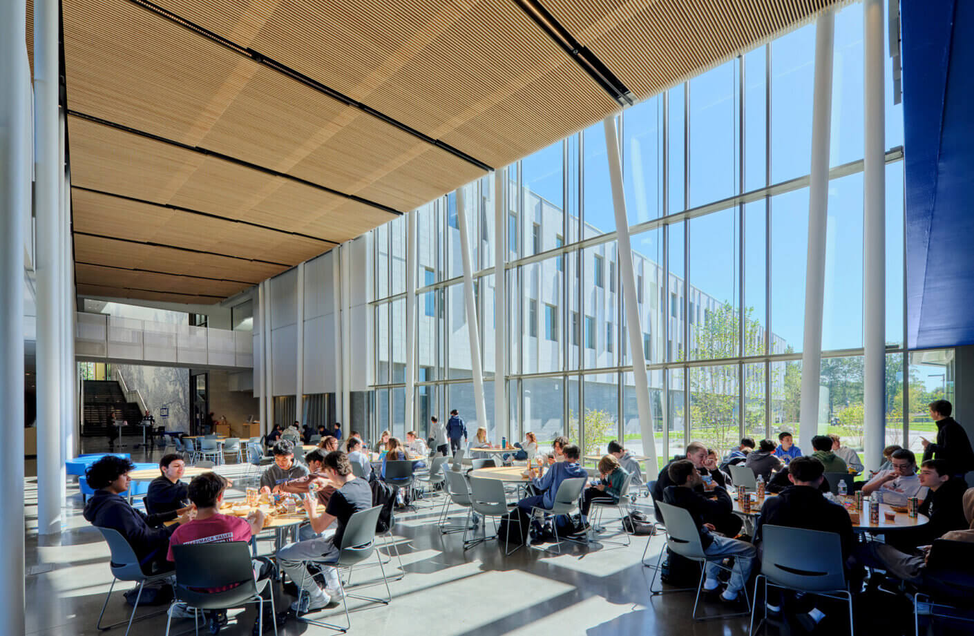 Bright, modern cafeteria with students dining and socializing around tables. Natural light floods in.