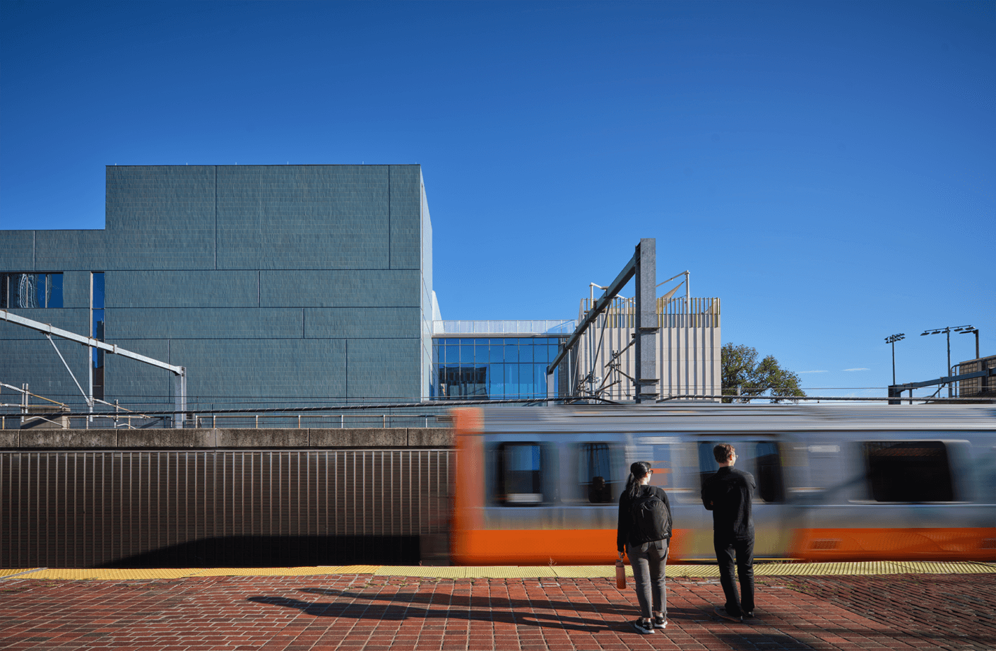 Two people waiting at a train station with a modern school building in the background.