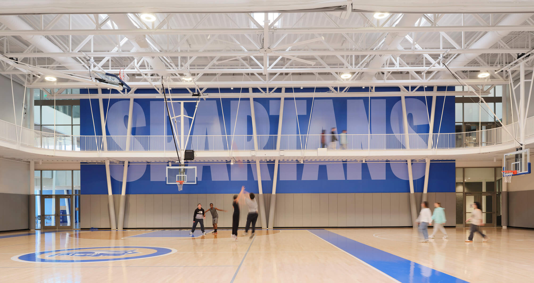 Indoor basketball court with SPARTANS mural, players shooting, and visitors walking.