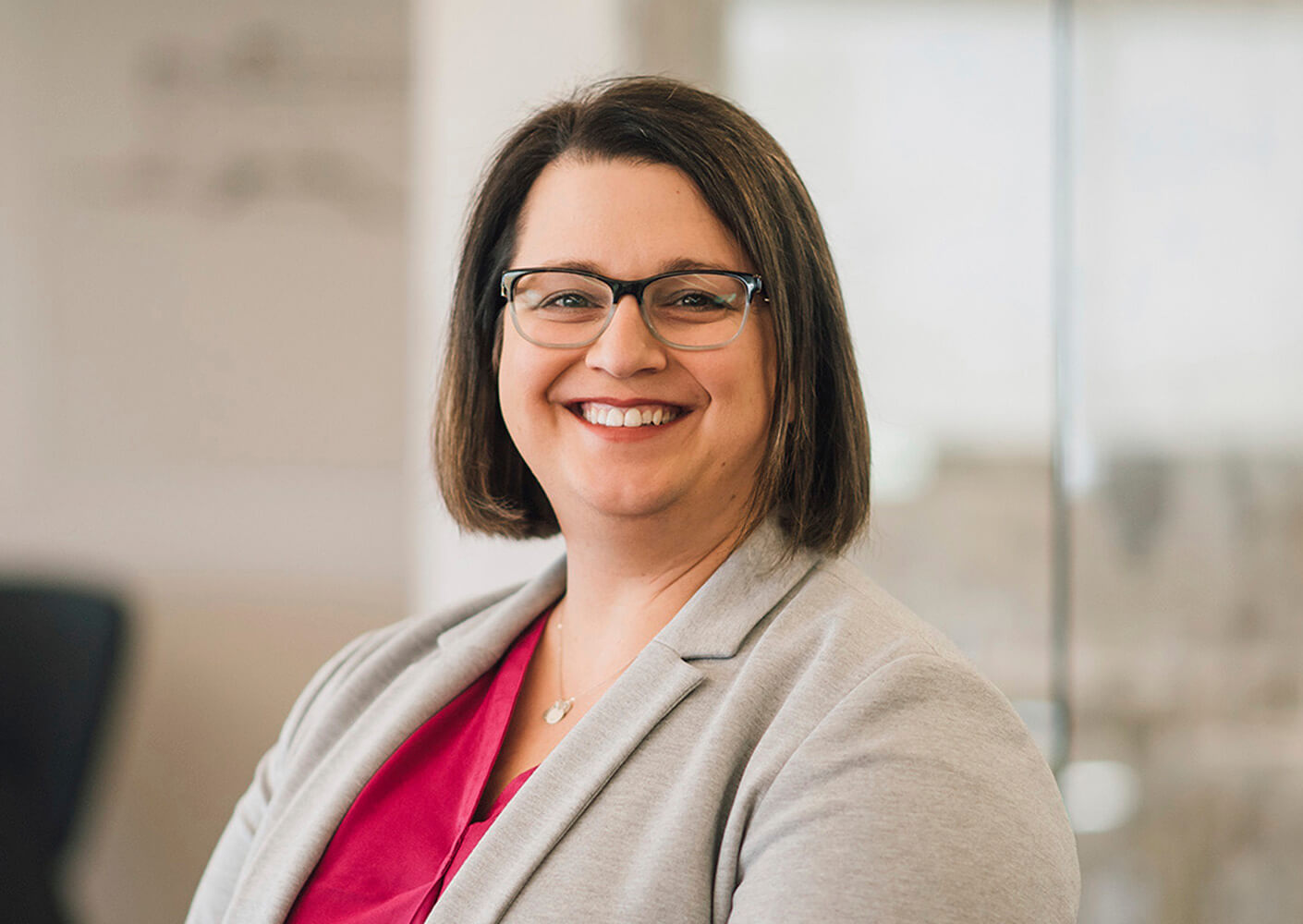 Professional woman smiling in an office setting, wearing a gray blazer and glasses.