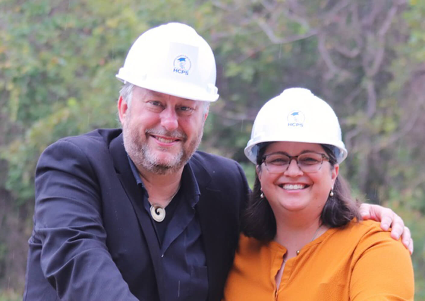 Two smiling professionals in hard hats pose together outdoors, showcasing teamwork and leadership.