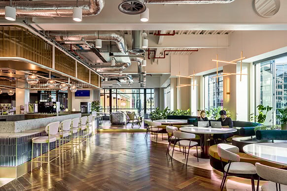 Bright open space featuring sleek lines, industrial elements, and varied seating arrangements, complemented by large windows and a herringbone wood floor.