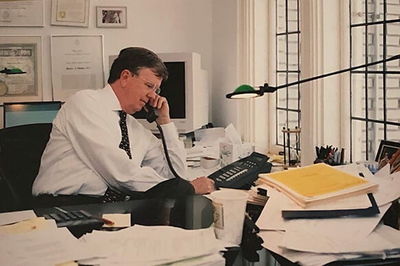 Henry Mann on the phone at a cluttered desk with papers and a computer. 