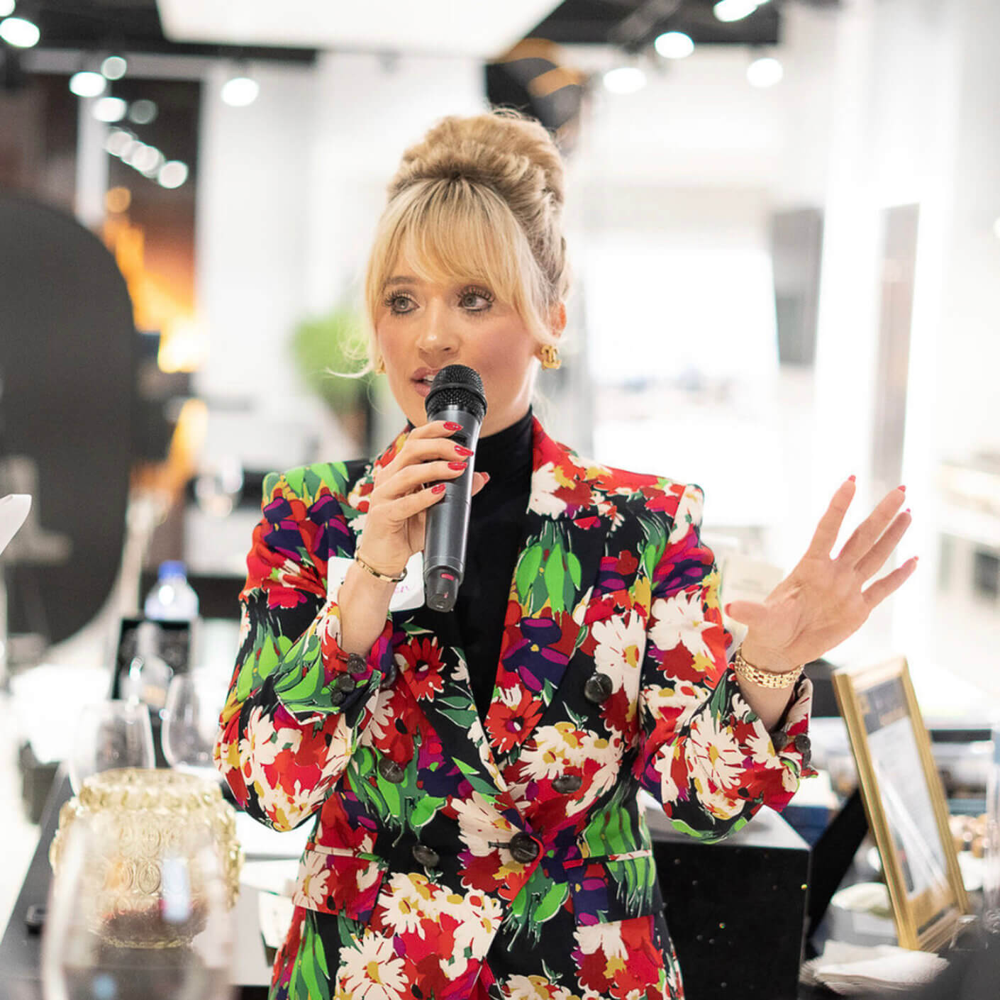 Andrea in a floral suit speaking into a microphone at an event, engaging the audience.