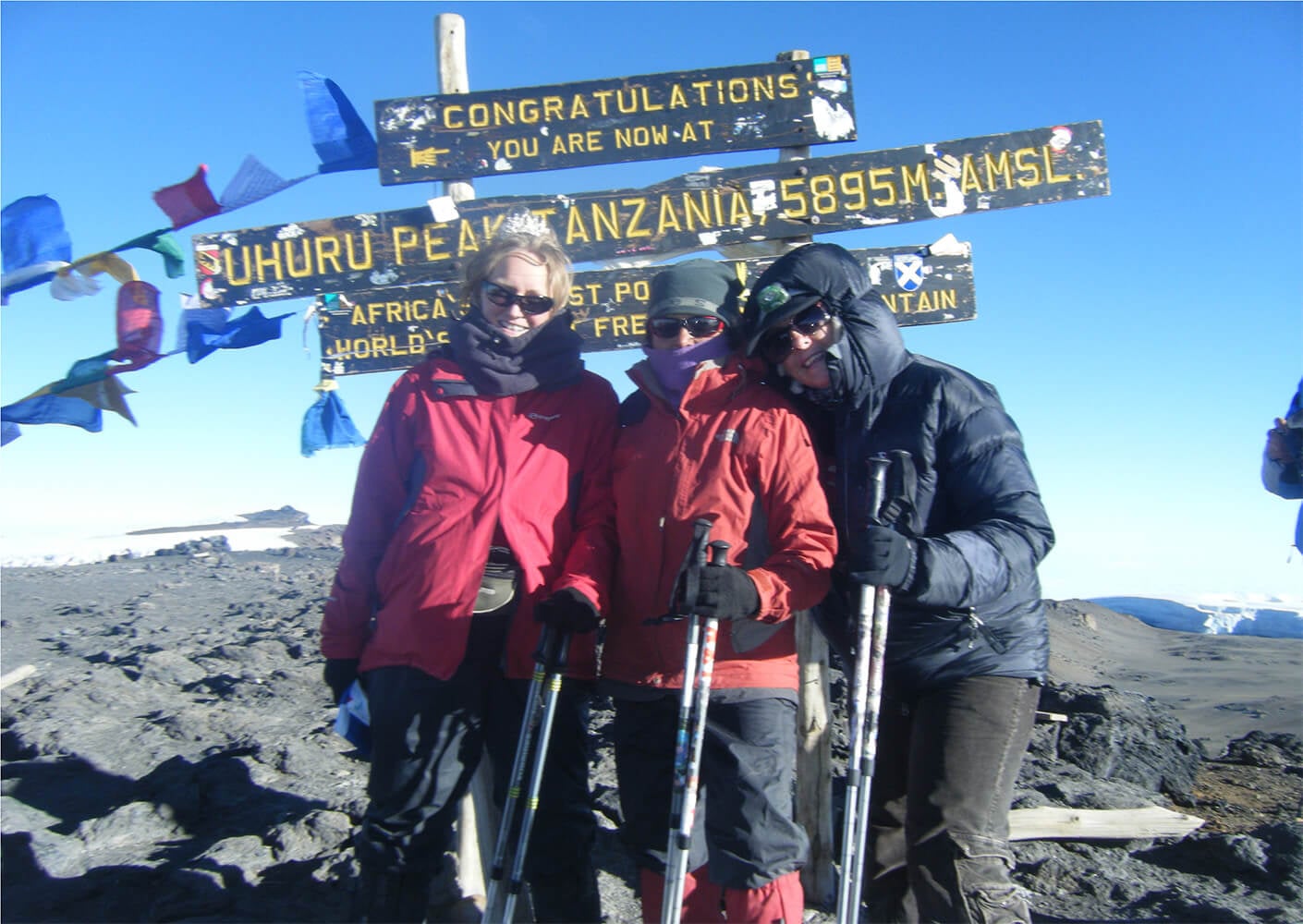 Three hikers celebrating at Uhuru Peak, Tanzania, with colorful prayer flags in the background.