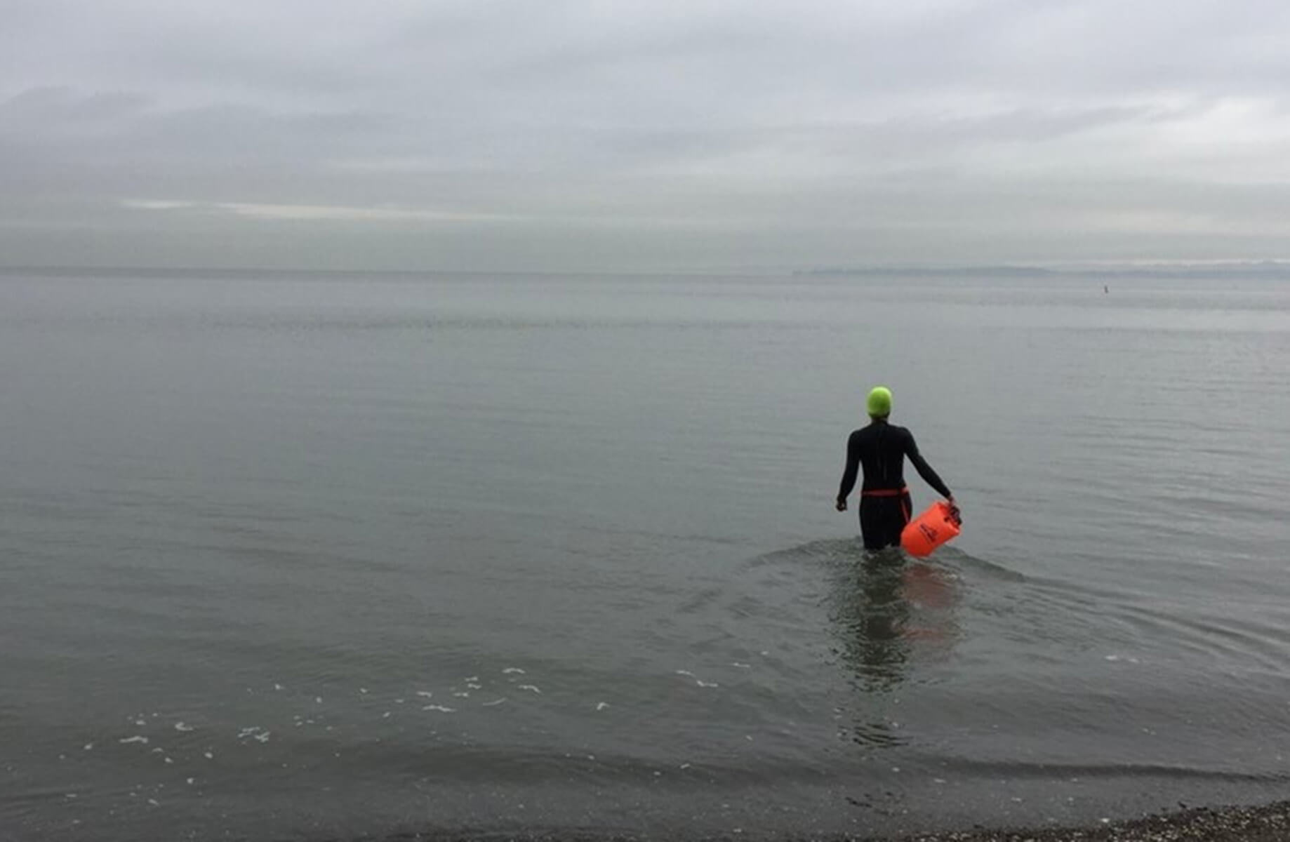 Swimmer in a wetsuit entering the calm water, holding a buoy, under a cloudy sky.