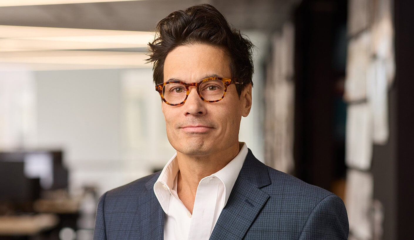 Professional man in glasses wearing a blazer, posing in a modern office setting.