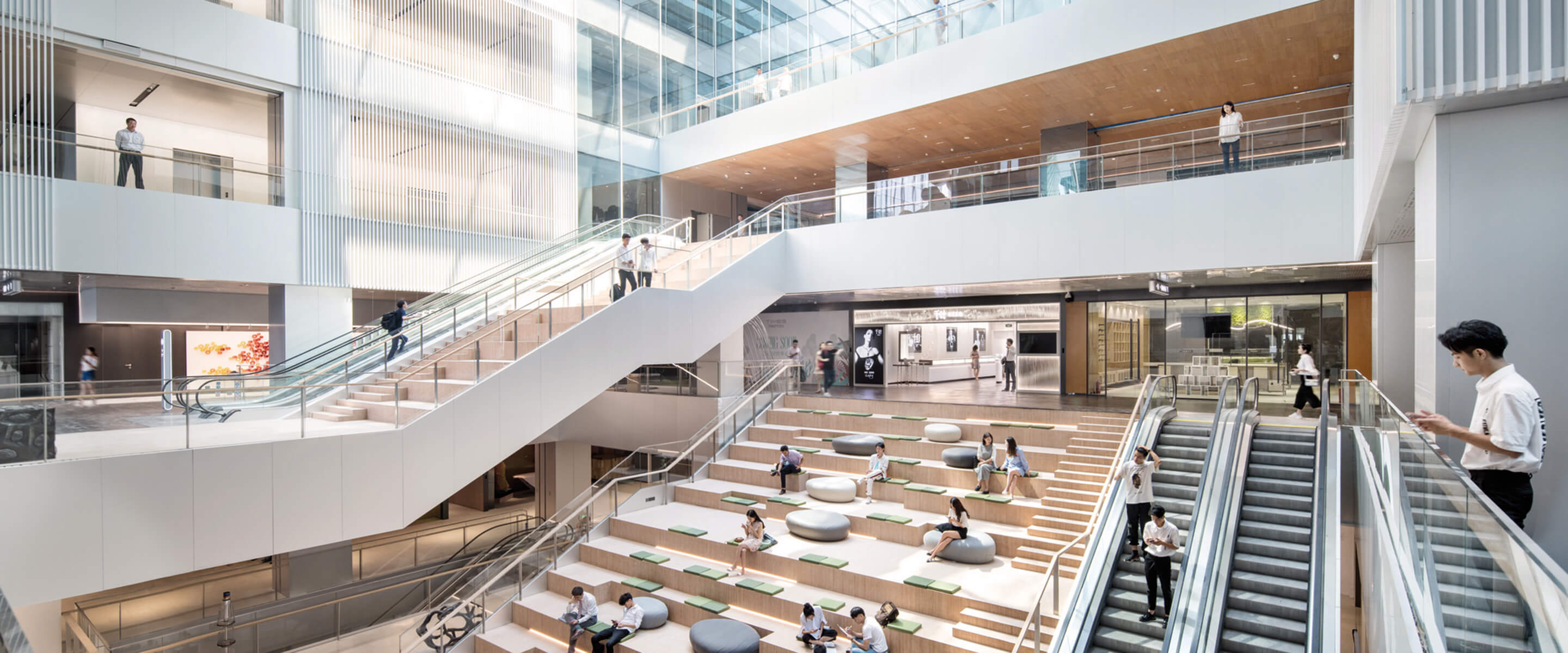 An expansive atrium featuring a blend of open spaces and multi-level seating areas. Natural light pours in through large glass panels, illuminating the wooden accents and sleek architectural lines. An elegant staircase winds through the space, connecting various levels, while comfortable seating invites visitors to relax. The interior emphasizes transparency and fluid movement, showcasing a dynamic layout that encourages social interaction and collaboration.