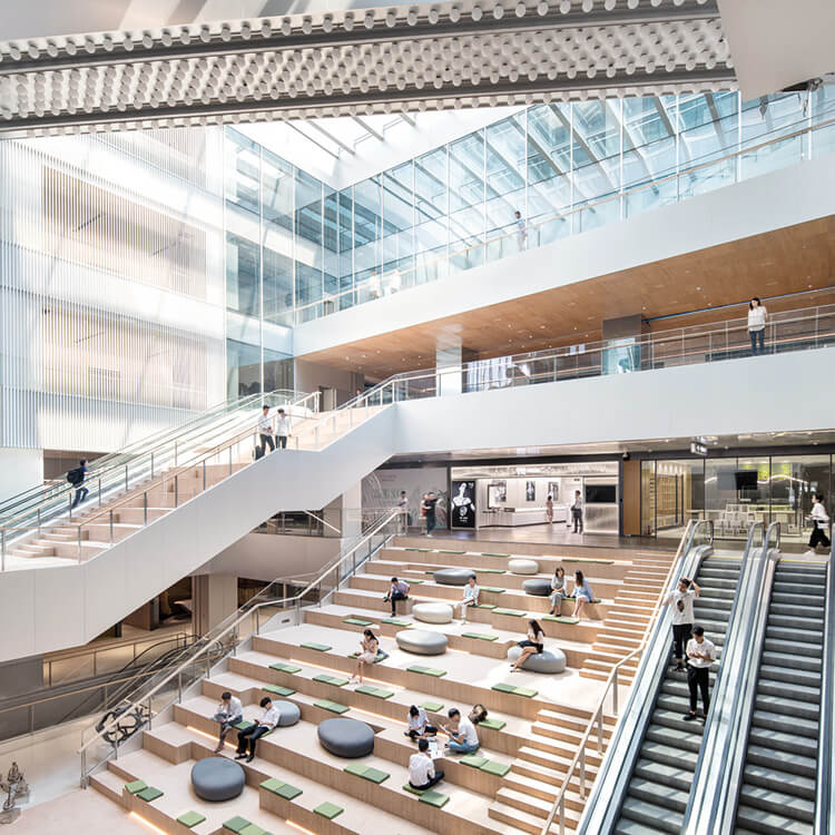 An expansive atrium featuring a blend of open spaces and multi-level seating areas. Natural light pours in through large glass panels, illuminating the wooden accents and sleek architectural lines. An elegant staircase winds through the space, connecting various levels, while comfortable seating invites visitors to relax. The interior emphasizes transparency and fluid movement, showcasing a dynamic layout that encourages social interaction and collaboration.