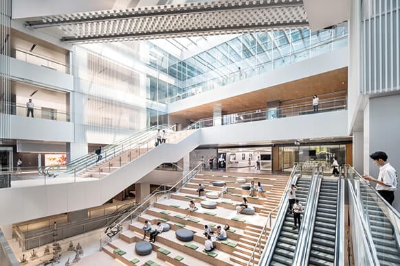 An expansive atrium featuring a blend of open spaces and multi-level seating areas. Natural light pours in through large glass panels, illuminating the wooden accents and sleek architectural lines. An elegant staircase winds through the space, connecting various levels, while comfortable seating invites visitors to relax. The interior emphasizes transparency and fluid movement, showcasing a dynamic layout that encourages social interaction and collaboration.