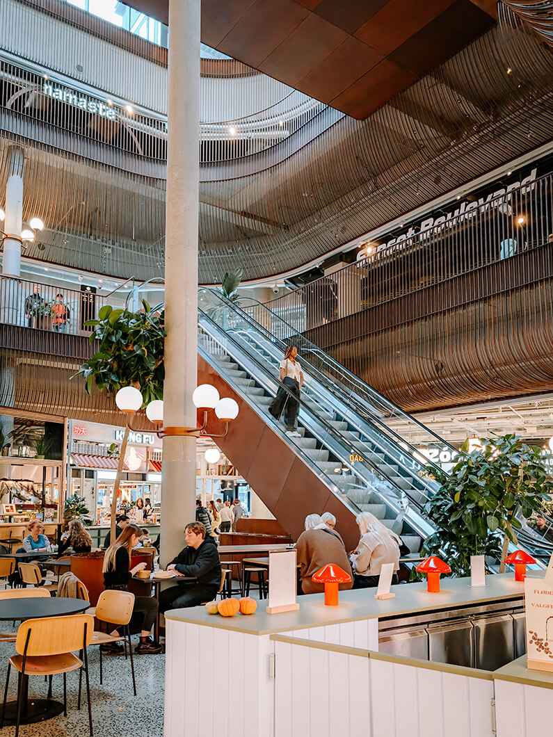 Modern interior of a bustling mall with escalators, plants, and cozy seating areas.
