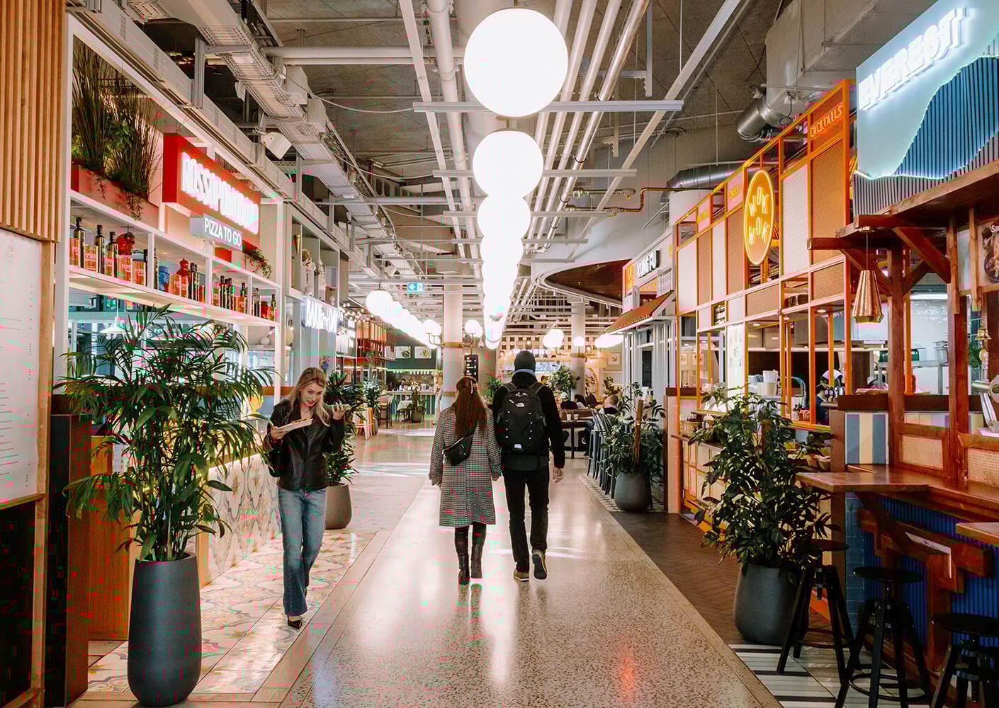 People walking through a vibrant food hall with various dining options and plants.