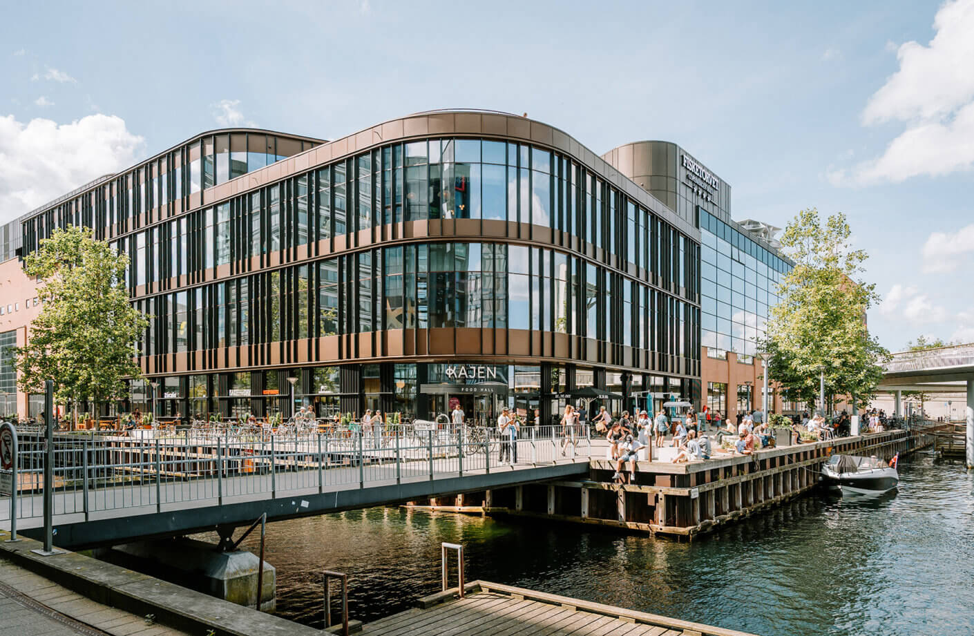 Modern waterfront building with people enjoying a food hall and relaxing by the canal.