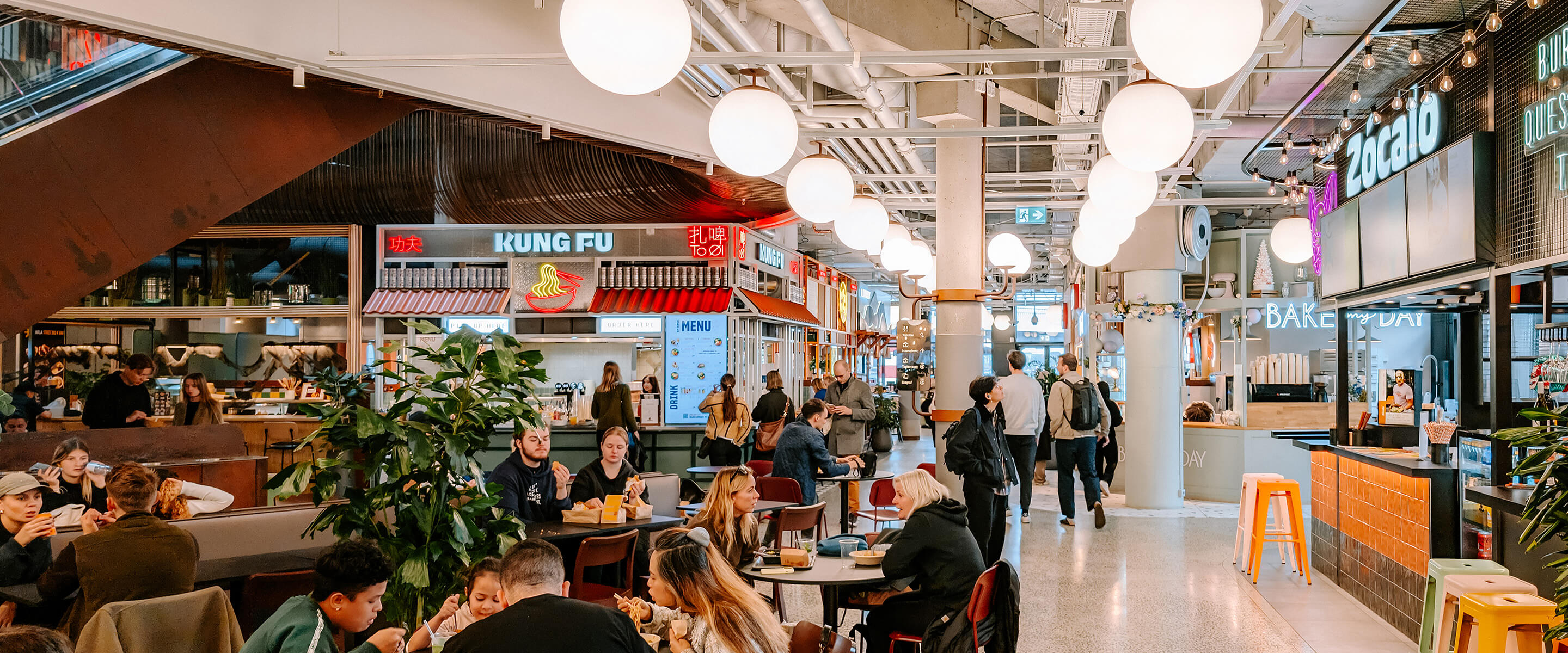 Busy food hall with diverse food stalls and patrons enjoying meals in a vibrant atmosphere.