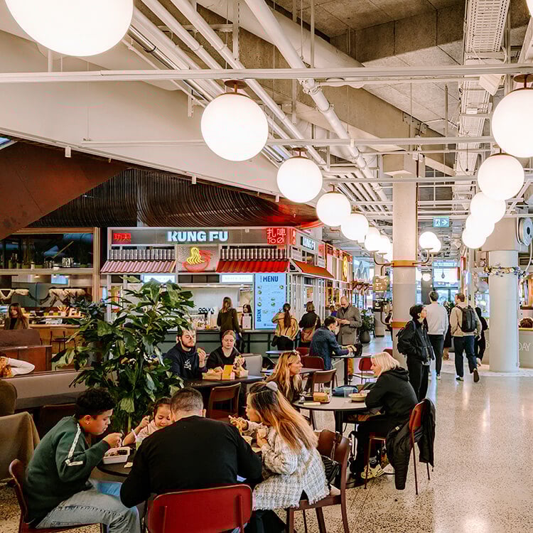 Busy food hall with diverse food stalls and patrons enjoying meals in a vibrant atmosphere.