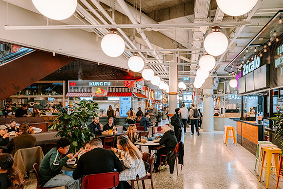 Vibrant food market bustling with diners and diverse food stalls under glowing lights.