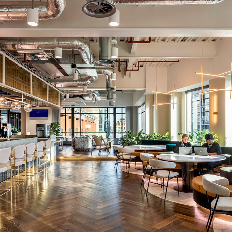 Bright open space featuring sleek lines, industrial elements, and varied seating arrangements, complemented by large windows and a herringbone wood floor.