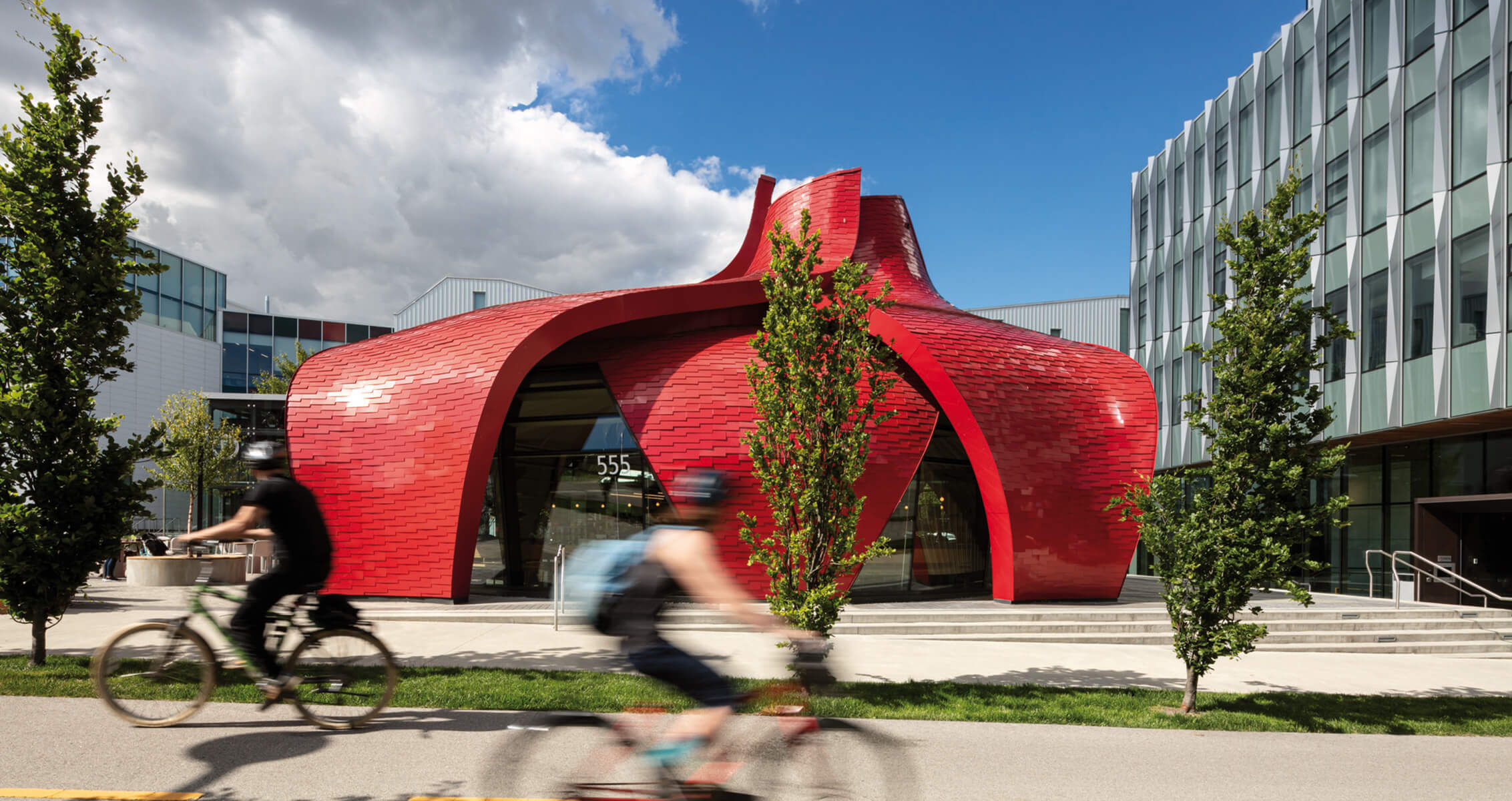 Vibrant red building with organic curves, featuring a distinctive tile facade and geometric glass elements, surrounded by greenery and cyclists.